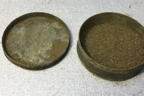 En gammal snusdosa med lock bredvid, fylld med utgånget snus.