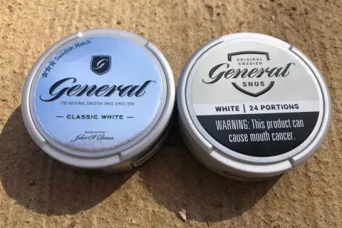 Två dosor General snus, en Classic White och en White, ligger på en sandig yta.