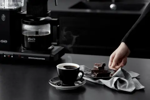 En hand sträcker sig efter chokladbitar bredvid en kopp ångande kaffe från en Moccamaster. Kanske är det dåligt kaffe, men chokladen ser god ut.