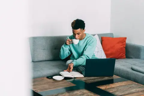 Ung man sitter i soffan med en kopp, kanske en mate drink, och läser en bok. En laptop står på bordet.