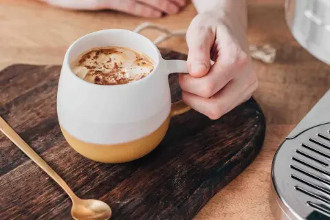 En hand håller en mugg med en krämig pumpa latte, toppad med kanel. En guldig sked ligger bredvid.