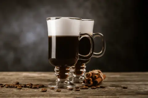 Två glas med kaffedrink Cointreau, toppade med grädde, står på ett träbord med kaffebönor och kanelstänger.