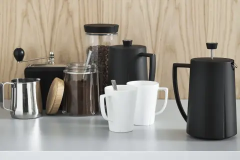 Kaffebönor och malet kaffe i glasburkar, en kaffekvarn, två vita muggar och två svarta presskannor. Perfekt för att utforska olika presskaffe sorter.