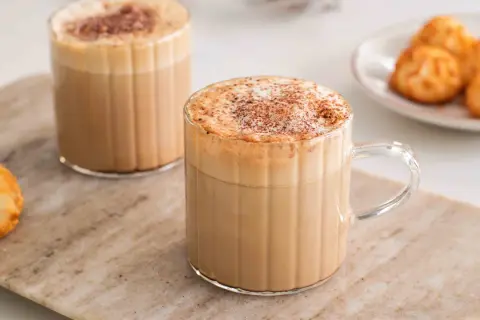 Två glas med krämigt skummande cafe americano, toppade med kanel. Bredvid står en liten tallrik med gyllene kokoskakor.