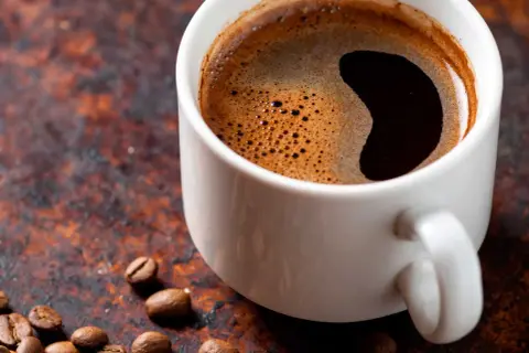 En vit kopp med starkt kaffe och skummig yta, omgiven av kaffebönor på en rustik bakgrund.