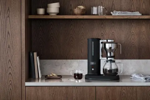 En stilren kaffebryggare i svart och krom, bredvid en glaskaraff med kaffe. På hyllan ovanför finns skålar och glas. En av flera olika kaffebryggare som passar in i ett modernt kök.