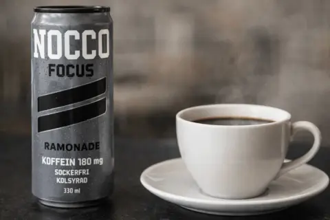En burk NOCCO Focus Ramonade med 180 mg koffein bredvid en ångande kopp kaffe. Perfekt för att kickstarta dagen med extra espresso koffein.