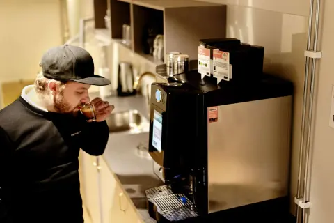 Man med keps njuter av en espresso i fikarummet på arbetsplatsen. En modern kaffemaskin står redo för nästa kopp.