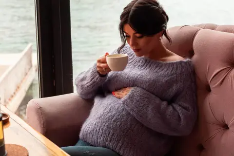Pregnant woman in a purple sweater holding a cup of tea, resting her hand on her belly.