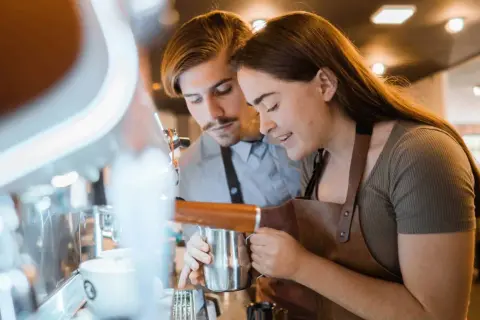 Två personer lär sig barista-yrket vid en espressomaskin. Hon håller en mjölkkanna, han tittar på.