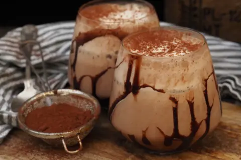 Två glas med krämig kaffe med choklad, garnerade med chokladsås och kakaopulver. En sil med kakaopulver står bredvid.