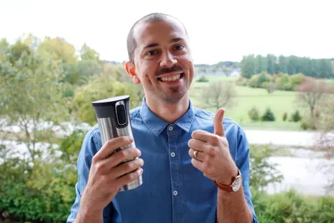 Glad man med tummen upp håller en termosmugg, redo för sitt fett kaffe utomhus.