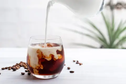 Mjölk hälls i ett glas med iskaffe på snabbkaffe. Kaffebönor ligger utspridda på bordet.