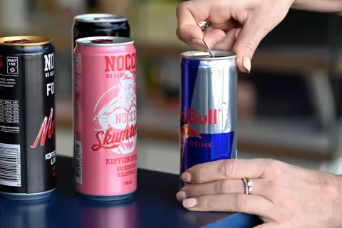 En hand öppnar en burk Red Bull, bredvid NOCCO-burkar. Koffein redbull för energi.