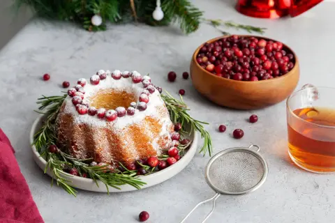 En vackert dekorerad sockerkaka med lingon och rosmarin, perfekt för julfika till många. En skål med lingon och en kopp te står redo.