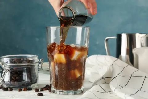En hand häller mörkt kaffe över is i ett glas. Bredvid står en burk med kaffebönor. Kaffe smakar surt om det inte är perfekt bryggt.