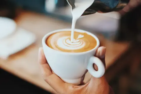 En hand håller en vit kaffekopp där mjölk hälls i för att skapa lattekonst.