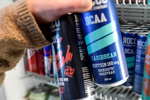 En hand tar en NOCCO Caribbean ur ett kylskåp fyllt med energidrycker. Frågan "hur många nocco per dag" dyker upp när man ser alla dessa.