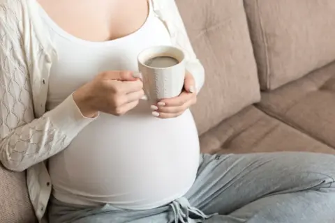 En gravid kvinna sitter på en soffa och håller en mugg med kaffe.