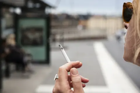 En kvinna röker en cigarett vid en busshållplats. Bilden kan illustrera prisutveckling cigaretter.
