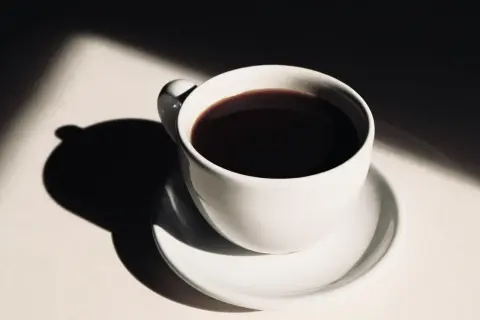 En vit kaffekopp med mörkt kaffe, belyst av solljus som skapar dramatiska skuggor. Hur påverkar kaffe blodtrycket?