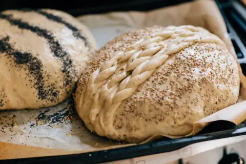Två nybakade bröd på bakplåtspapper, ett toppat med vallmofrön och ett med sesamfrön och flätat mönster. Perfekta fika idéer!