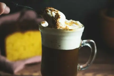En sked med vispad grädde och kanel toppar en varm kaffe Karlsson utan Baileys. I bakgrunden syns en gul kaka.
