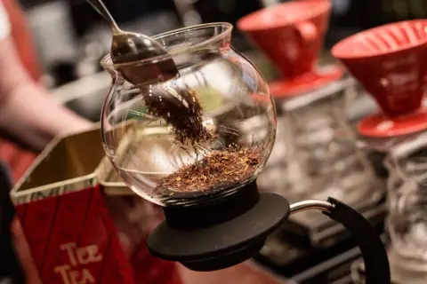 En sked lägger teblad i en glaskaraff, redo för att brygga te utan koffein. Röda kaffefilter står i bakgrunden.