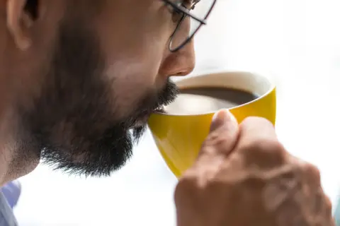 Man med glasögon dricker kaffe ur en gul mugg. Koffein ger energi, motverkar adenosin.