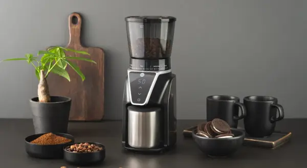 En modern kaffekvarn med digital display, fylld med bönor, bredvid skålar med malet kaffe och hela bönor. Två svarta muggar och en liten krukväxt fulländar scenen.