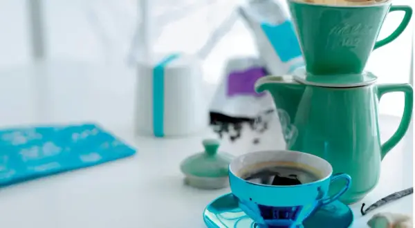 En turkos kaffekopp med fat står framför en grön kaffekanna med filterhållare. Kaffebönor och en vaniljstång ligger bredvid. En fin morgon med gott kaffe.