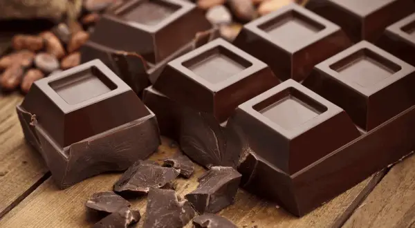 Mörk chokladbitar på träbord, med kakaobönor i bakgrunden. En härlig källa till kakao och koffein för en energikick.