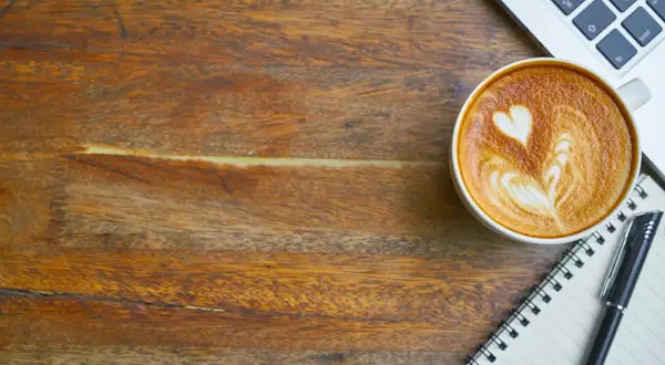 En kopp kaffe med lattekonst, en laptop och en anteckningsbok på ett träbord. Perfekt för att få lite koffein och inspiration.