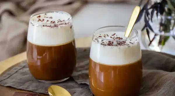 Två glas med kaffe drinkar med alkohol, toppade med grädde och chokladströssel. En gyllene sked vilar i det ena glaset.