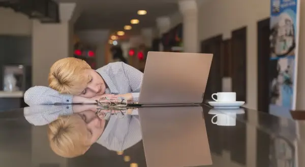 Kvinnan med blont hår sover vid ett bord med en laptop och en kaffekopp. Hon verkar trött, trots att hon inte blir pigg av kaffe.