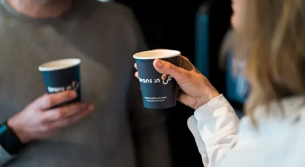 Två personer håller kaffemuggar. En kvinna håller en mugg med texten "Beans in Up". Kanske är det här svaret på vilket land dricker mest kaffe.