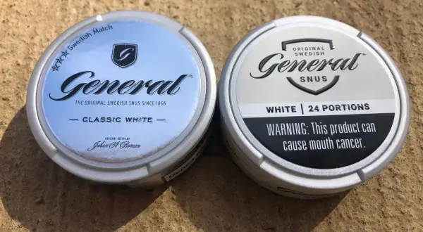 Två dosor General snus, en Classic White och en White, ligger på en sandig yta.