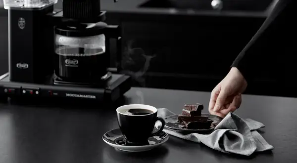 En hand sträcker sig efter chokladbitar bredvid en kopp ångande kaffe från en Moccamaster. Kanske är det dåligt kaffe, men chokladen ser god ut.