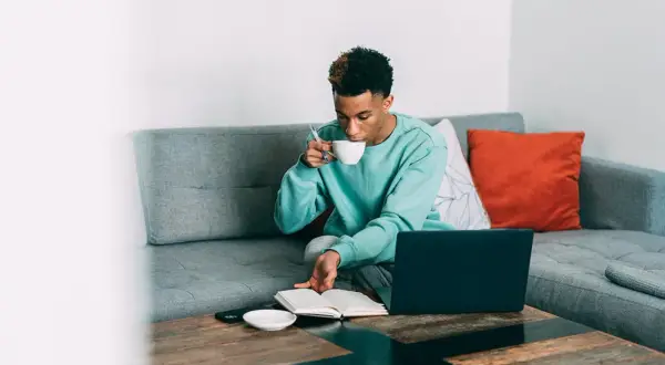 Ung man sitter i soffan med en kopp, kanske en mate drink, och läser en bok. En laptop står på bordet.