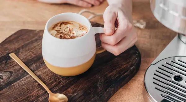 En hand håller en mugg med en krämig pumpa latte, toppad med kanel. En guldig sked ligger bredvid.