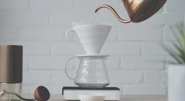 Vatten hälls från en kopparkanna över ett filter i en vit V60-bryggare, som står på en glaskaraff. En kaffekvarn och en skopa ligger bredvid. Detta visar hur brygger man kaffe.