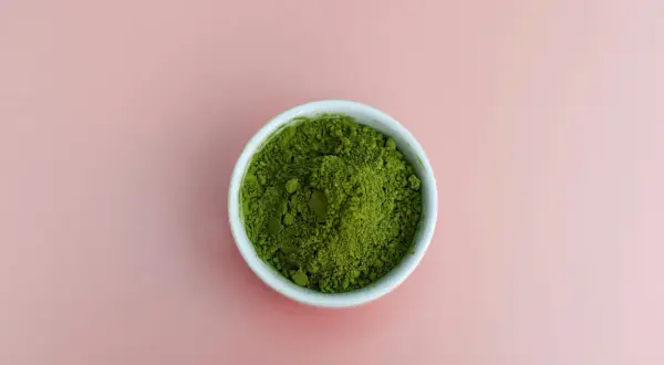 En skål med intensivt grönt matchapulver på en mjukrosa bakgrund. Undrar du vad smakar matcha? Det är en unik, lite bitter och gräsig smak.
