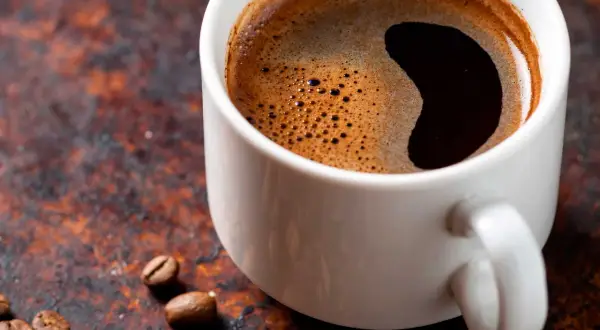 En vit kopp med starkt kaffe och skummig yta, omgiven av kaffebönor på en rustik bakgrund.