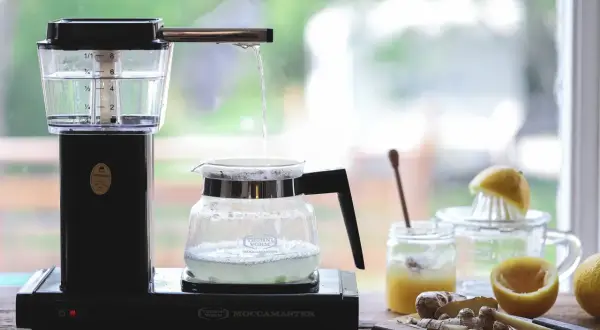 Vatten droppar från en kaffebryggare ner i en glaskanna, redo att brygga te. Ingredienser som citron och ingefära syns i bakgrunden.