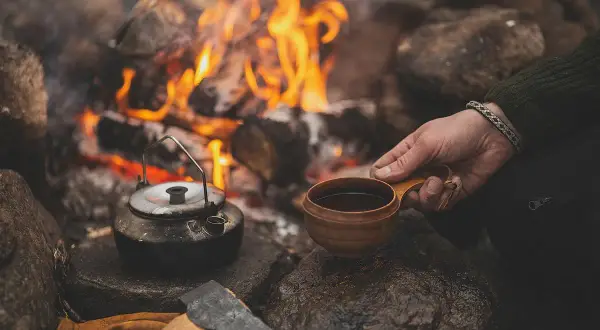 Vid en sprakande eld, med en kaffepanna och en träkopp, kan man brygga kokkaffe. En yxa och handskar ligger bredvid.