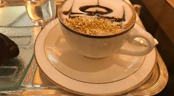 En lyxig kaffekopp med guldfoliering och chokladdrizzle, kanske det dyraste kaffet någonsin. Serveras på ett silverfat med ett glas vatten.