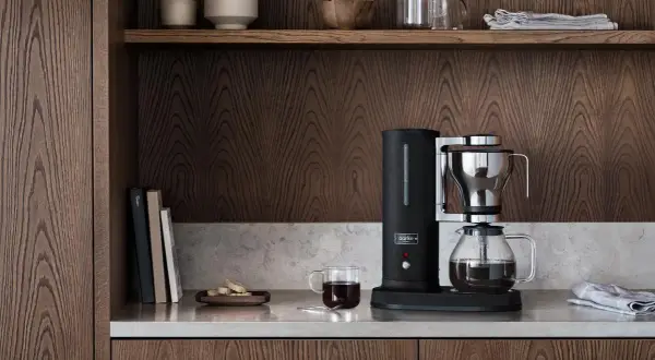 En stilren kaffebryggare i svart och krom, bredvid en glaskaraff med kaffe. På hyllan ovanför finns skålar och glas. En av flera olika kaffebryggare som passar in i ett modernt kök.