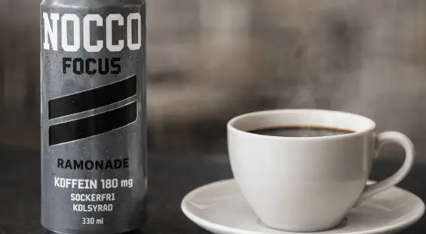 En burk NOCCO Focus Ramonade med 180 mg koffein bredvid en ångande kopp kaffe. Perfekt för att kickstarta dagen med extra espresso koffein.