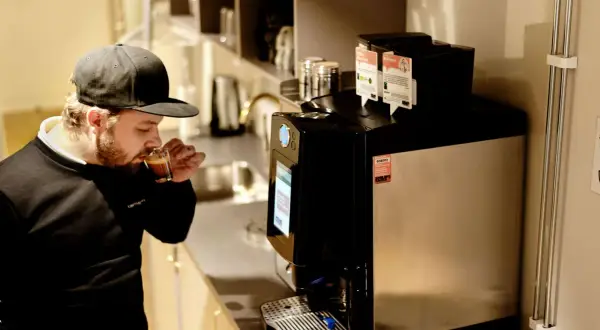 Man med keps njuter av en espresso i fikarummet på arbetsplatsen. En modern kaffemaskin står redo för nästa kopp.