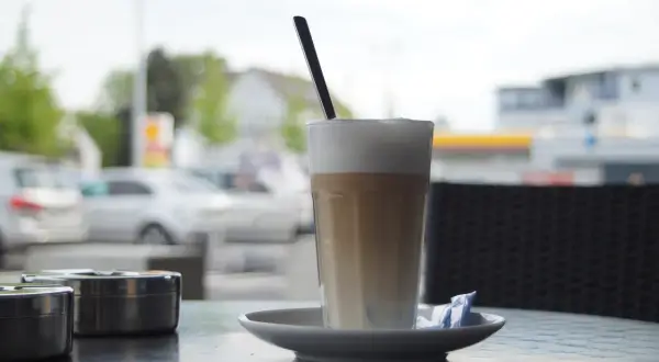 En hög latte macchiato med skummig mjölk och en sked, redo att avnjutas vid ett cafébord utomhus.